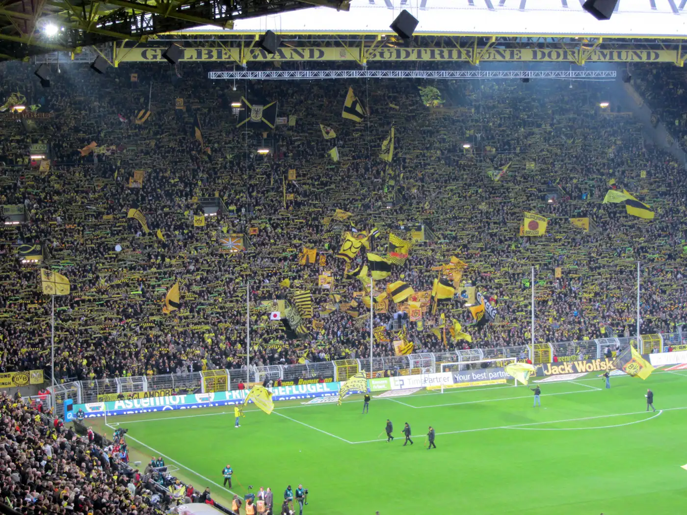 Grada de pie abarrotada en el Signal Iduna Park de Dortmund, con miles de aficionados ondeando banderas amarillas y negras bajo un cartel que dice «Gelbe Wand Südtribüne Dortmund», junto al césped iluminado.