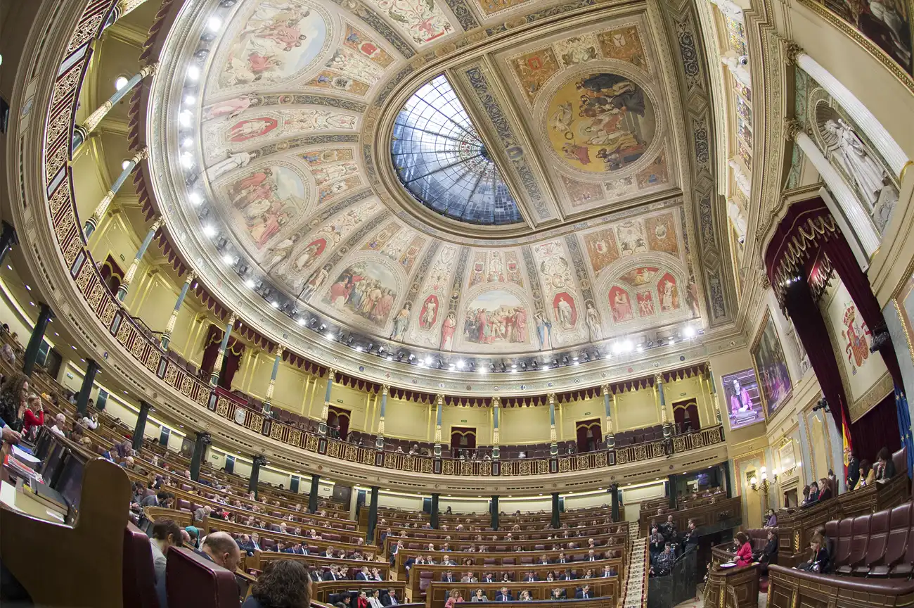 Vista general del hemiciclo del Congreso de los Diputados durante un pleno, tomada con objetivo muy angular: la cúpula de cristal y el techo de frescos dorados ocupan la mitad superior, debajo se ven las tribunas de madera y, en el nivel inferior, decenas de diputados sentados en los escaños; a la derecha, el dosel granate con el escudo de España que preside la tribuna.