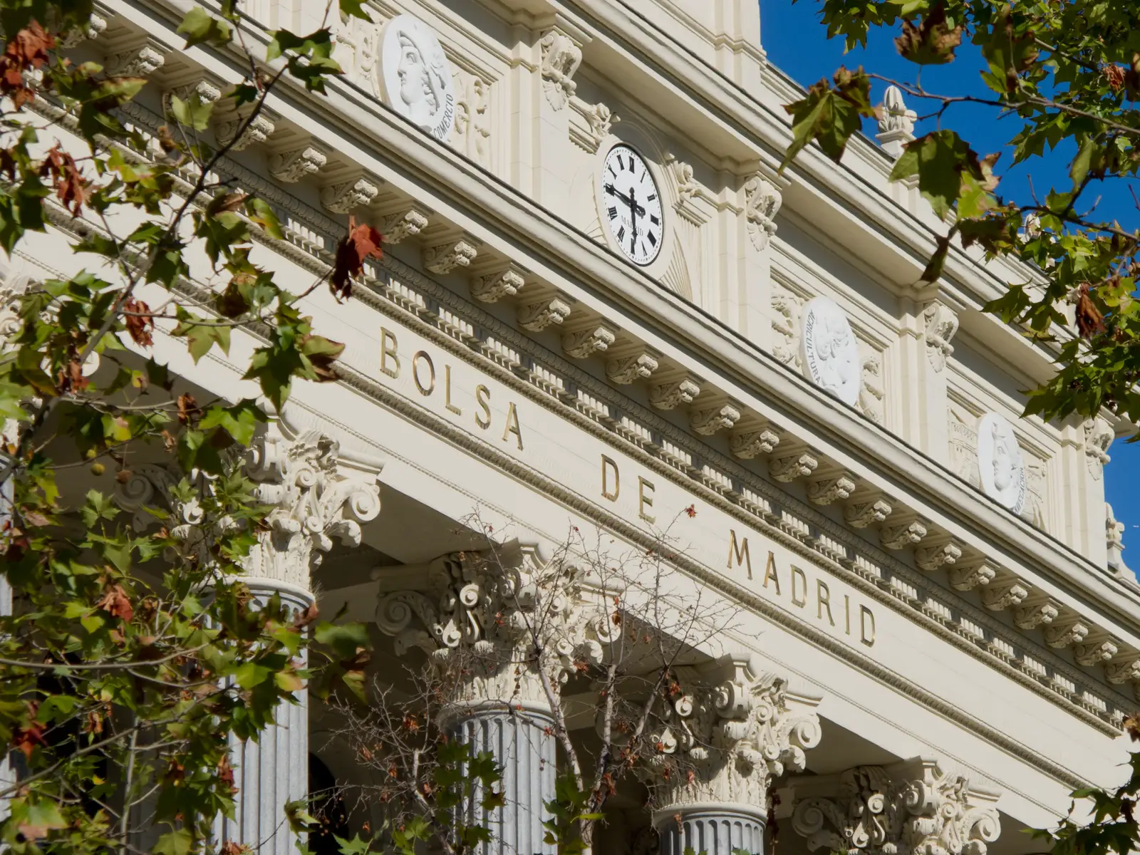 Fachada del Palacio de la Bolsa de Madrid vista desde abajo entre las ramas de un árbol: friso de piedra clara con las letras doradas «BOLSA DE MADRID», un reloj de esfera blanca sobre la cornisa, medallones con bustos y capiteles corintios recortados contra un cielo azul.