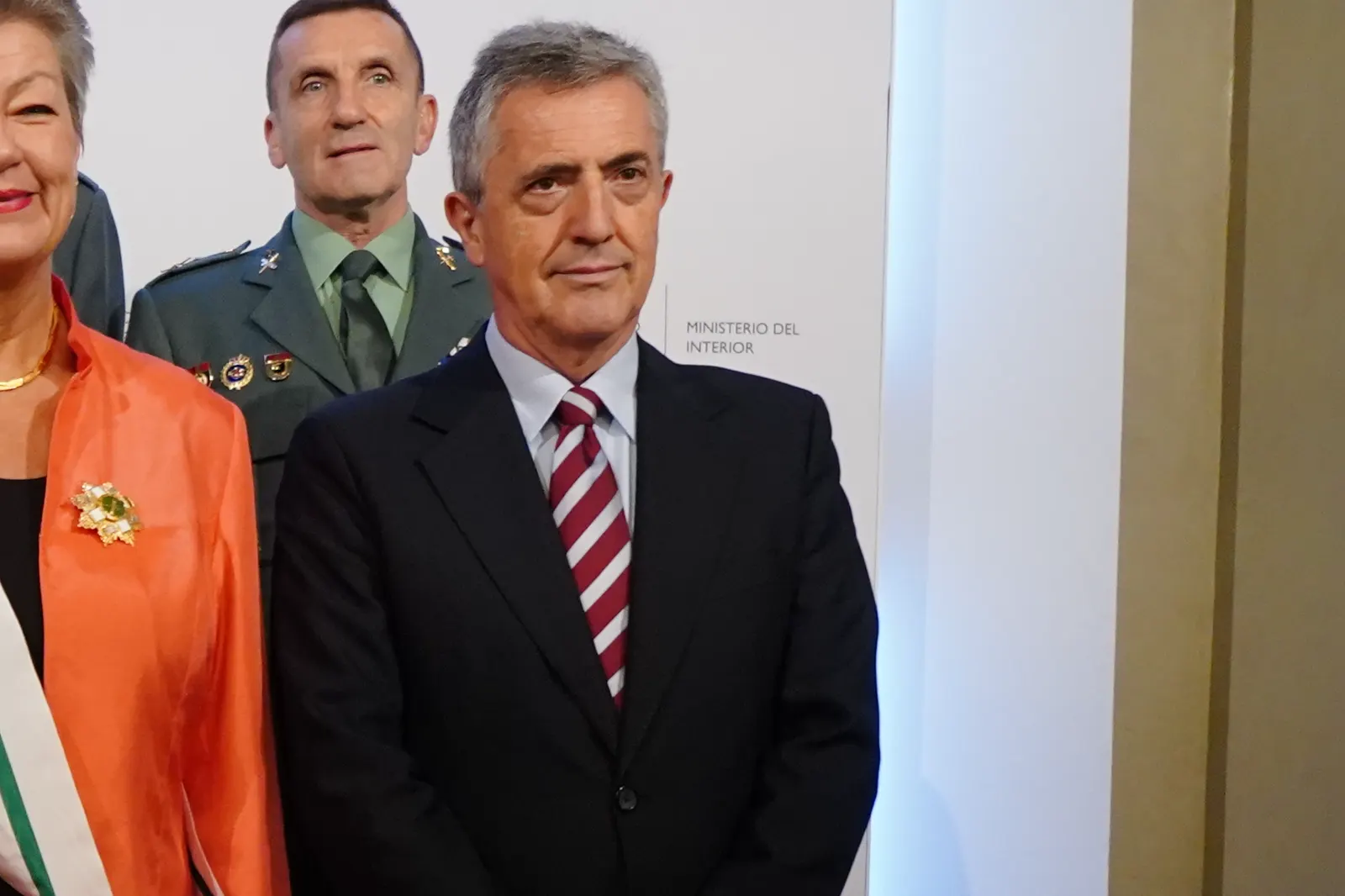 Leonardo Marcos, con traje oscuro y corbata de rayas granates y blancas, posa ante un panel con el rótulo «Ministerio del Interior»; detrás, un mando de la Guardia Civil de uniforme y, a su lado, una mujer con chaqueta naranja y banda de condecoración.