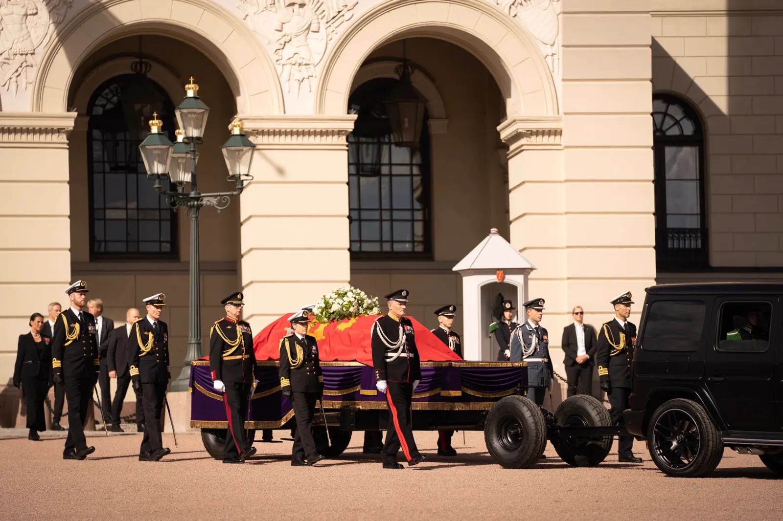 El féretro del rey Harald V, cubierto por el estandarte real rojo con el león dorado y una corona de flores blancas, avanza sobre una cureña con faldón morado remolcada por un todoterreno negro frente a la fachada del Palacio Real de Oslo, flanqueado por oficiales de uniforme de gala.