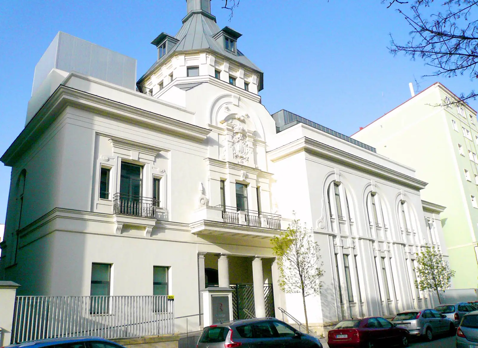 Edificio blanco de dos plantas con torreón, balcones de forja y escudo labrado sobre la puerta principal, con coches aparcados delante y el cielo despejado al fondo.