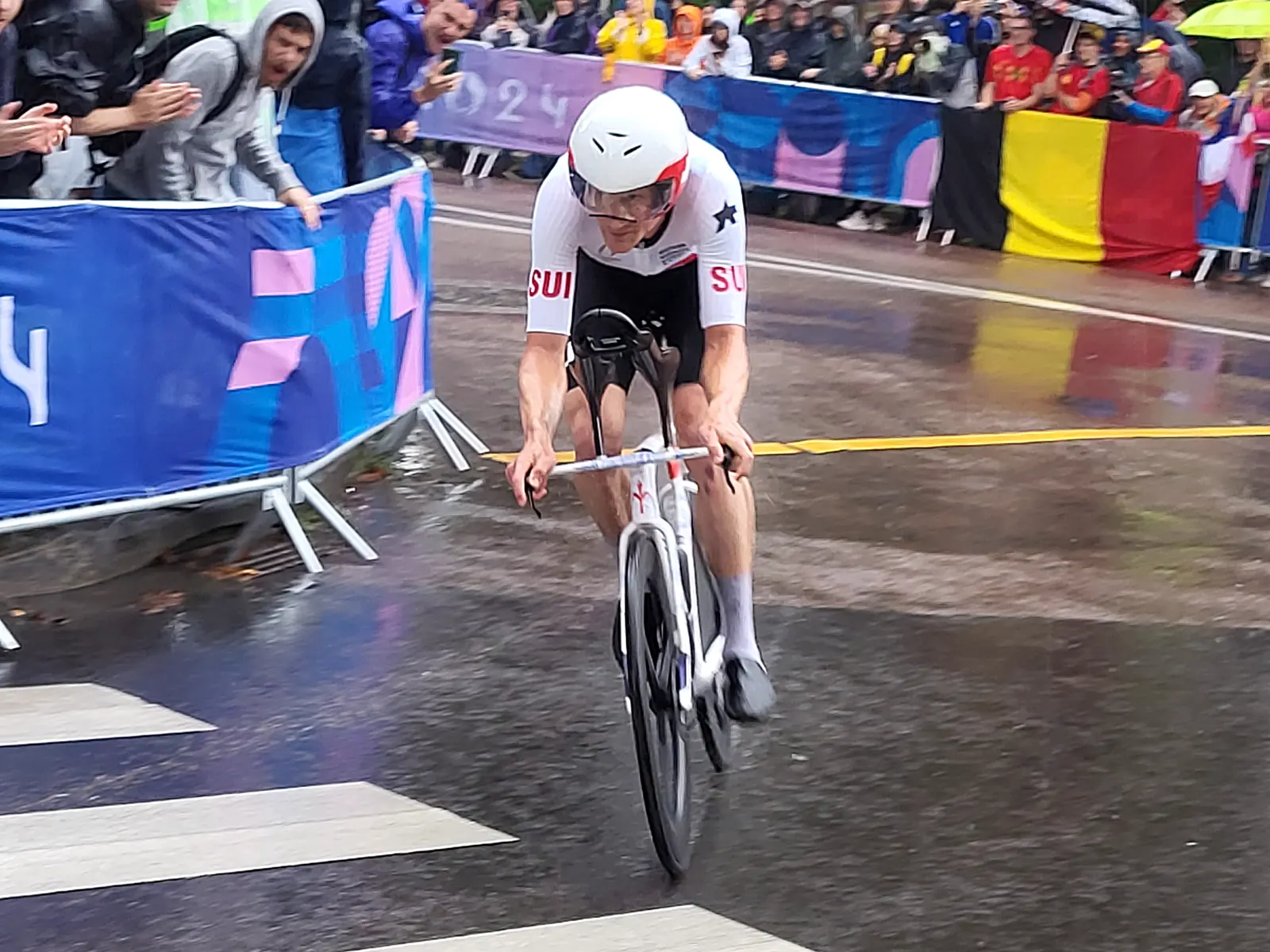 Un ciclista con maillot blanco y rojo de Suiza y casco aerodinámico pedalea agachado sobre una bicicleta de contrarreloj por una carretera mojada, entre vallas y público que aplaude bajo la lluvia.