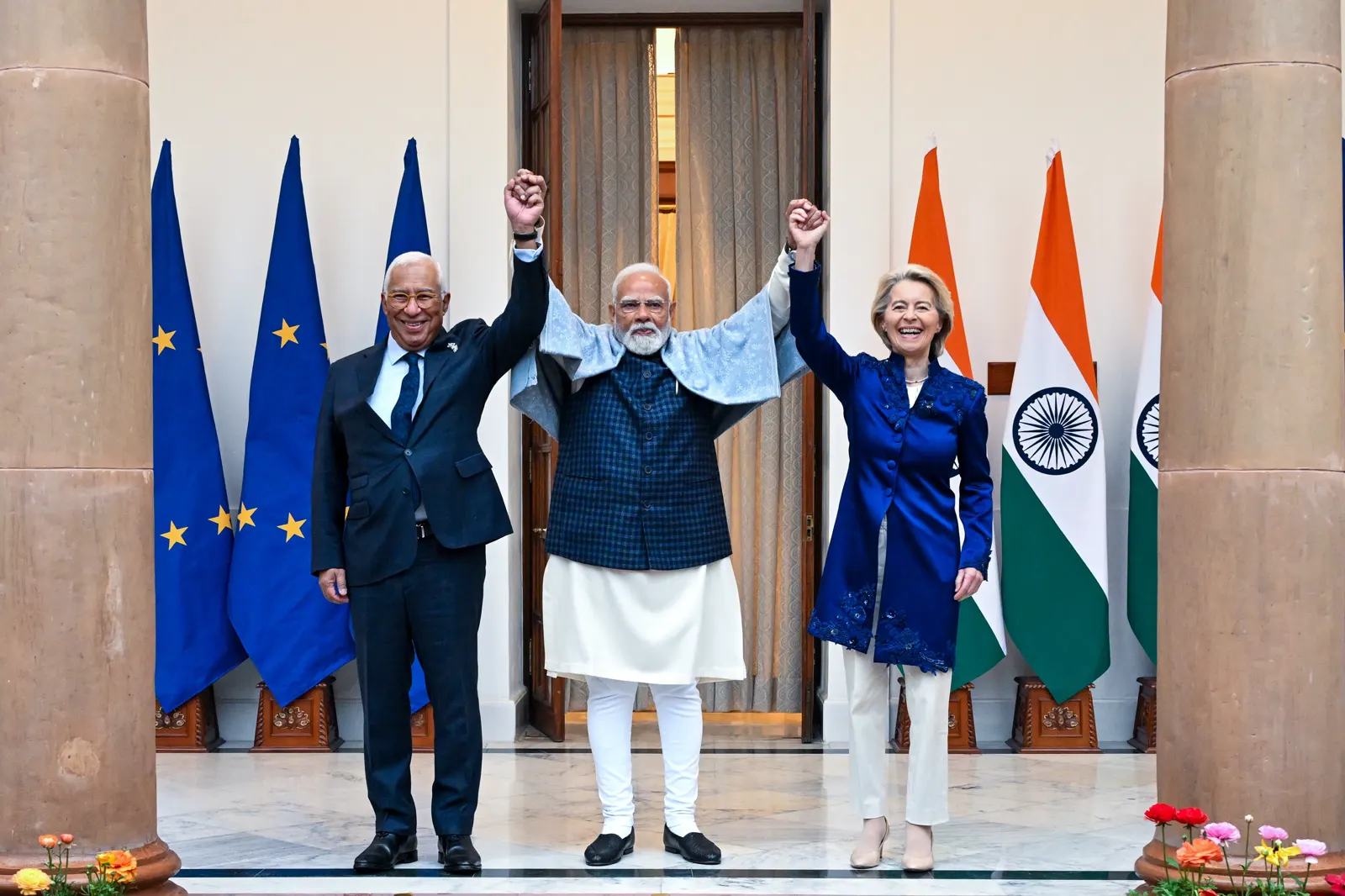 António Costa, Narendra Modi y Ursula von der Leyen, de pie ante banderas de la Unión Europea e India, sonríen con los brazos levantados y las manos entrelazadas.
