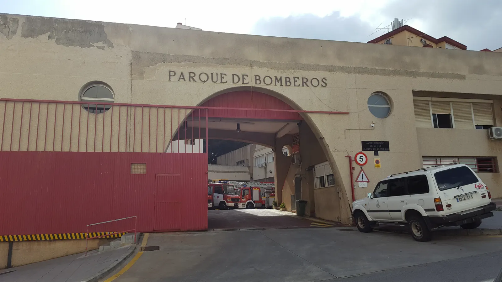 Fachada de un parque de bomberos con el rótulo «Parque de Bomberos» sobre un gran arco de entrada; al fondo, dentro de la nave, se ven dos camiones de bomberos rojos aparcados, y a la derecha un todoterreno blanco circula por la calle.