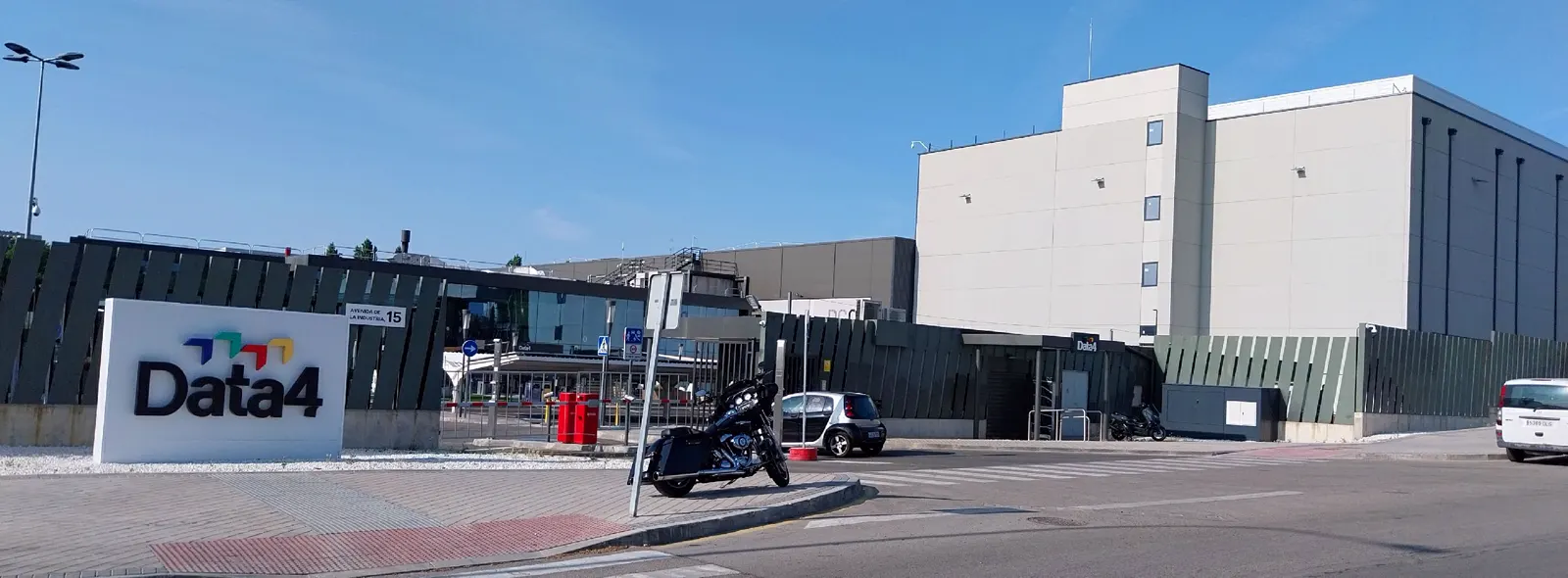 Entrada de un campus industrial con un cartel blanco con el logotipo Data4, naves sin ventanas tras una valla oscura, una moto y varios coches aparcados