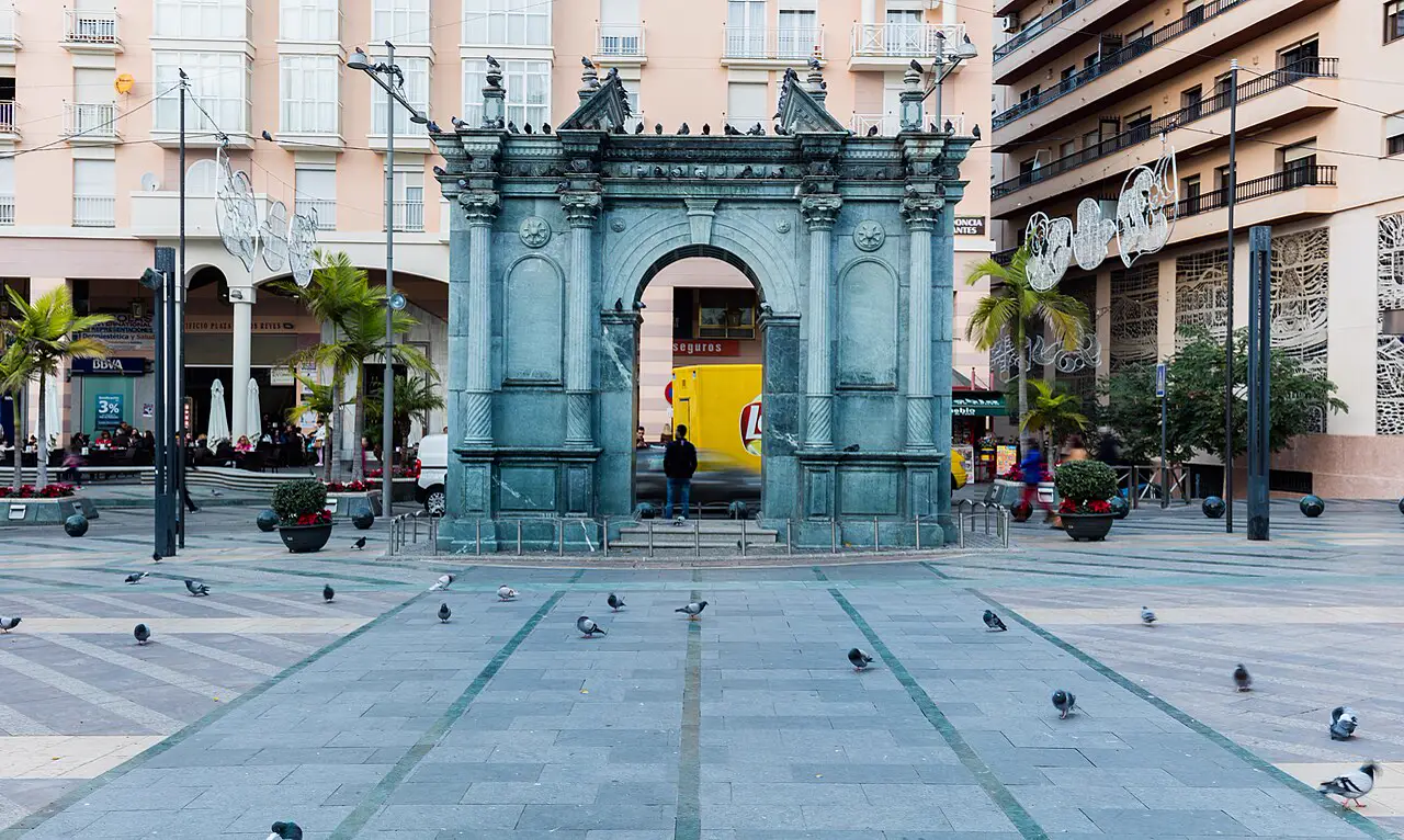 Gran portada monumental de piedra verdosa, con columnas y un arco central, aislada en medio de una plaza empedrada por la que caminan palomas; al fondo, bloques de viviendas con balcones, palmeras y farolas.