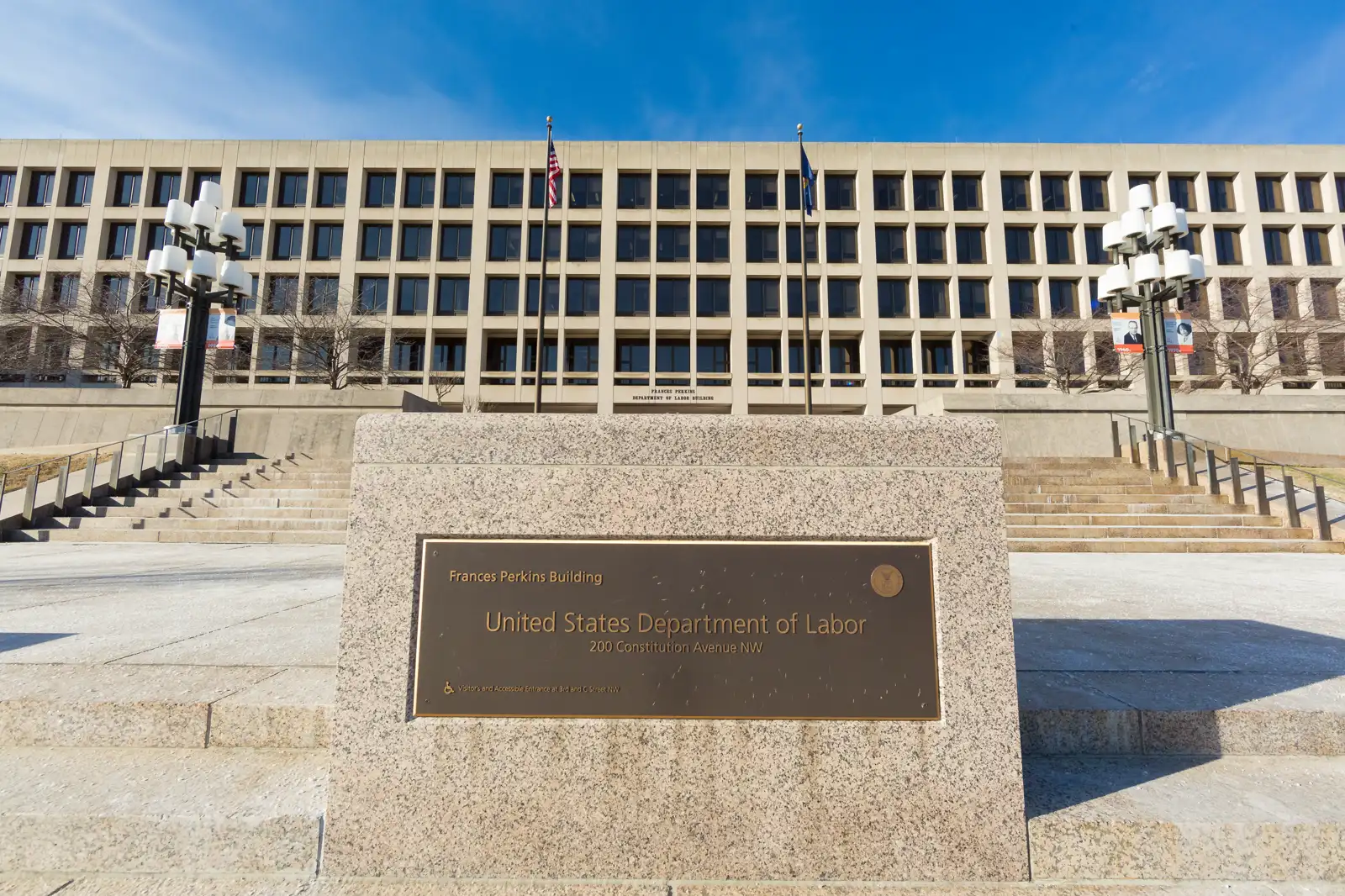 Monolito de granito con una placa de bronce que dice «Frances Perkins Building. United States Department of Labor. 200 Constitution Avenue NW», ante la fachada de hormigón y la escalinata del edificio, con la bandera de Estados Unidos y cielo azul al fondo.