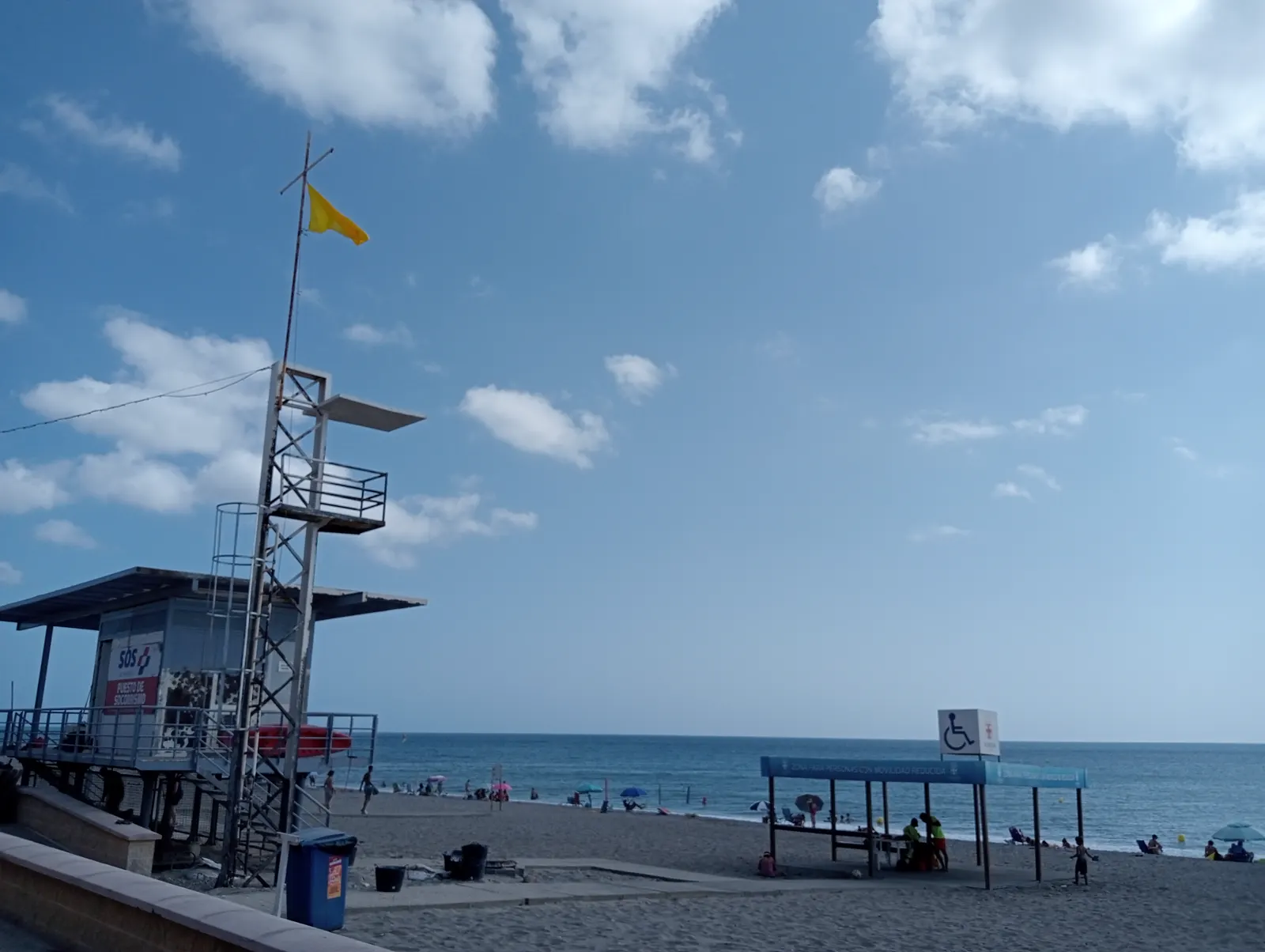 Puesto de socorrismo elevado en una playa española, con bandera amarilla izada en el mástil y bañistas a lo lejos junto a la orilla.