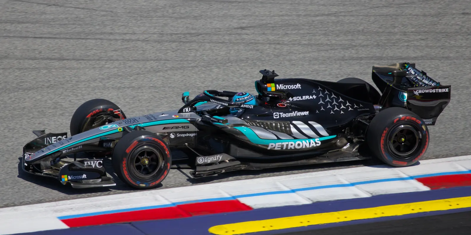 Monoplaza de Fórmula 1 de Mercedes, negro con franjas turquesa, rodando sobre el asfalto junto a un pianista rojo, blanco y azul