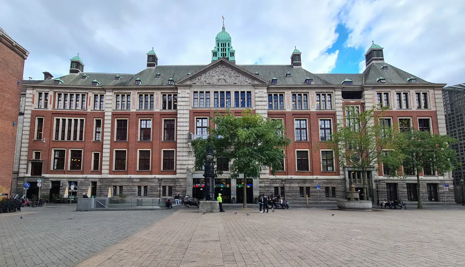 Fachada de ladrillo rojo y piedra del edificio de la bolsa de Ámsterdam, con torreones de cubierta verde, vista desde la plaza empedrada en la que hay transeúntes sentados y la escultura de un toro junto a la entrada.