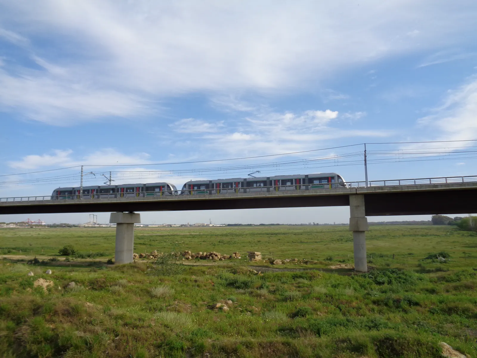 Un tren doble del metro de Sevilla circula por un viaducto sobre una llanura verde bajo un cielo con nubes