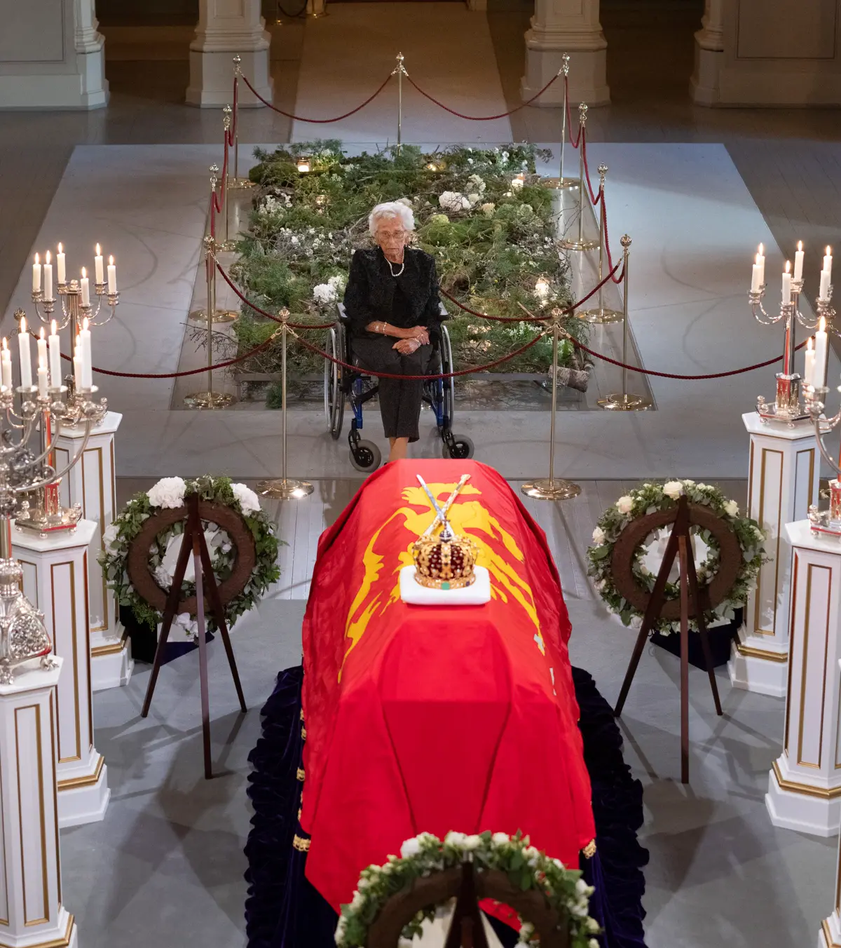 Una mujer mayor vestida de negro, sentada en silla de ruedas, permanece sola ante el féretro de Harald V, cubierto con el estandarte real rojo y con la corona encima, en la capilla del Palacio Real de Oslo, entre candelabros y coronas de flores.