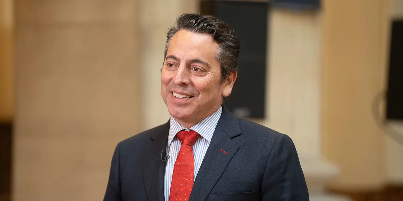 Hombre de mediana edad con traje azul marino, camisa de rayas y corbata roja, sonriente y con micrófono de solapa, en un interior de paredes claras desenfocado.