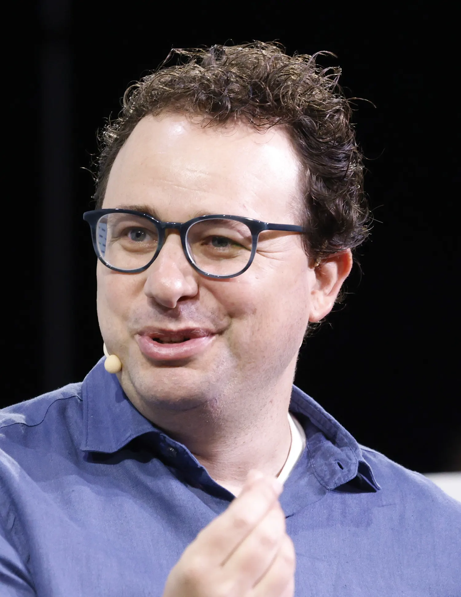 Retrato de Dario Amodei, con gafas oscuras de pasta, camisa azul y micrófono de diadema, hablando sobre un escenario con fondo negro.