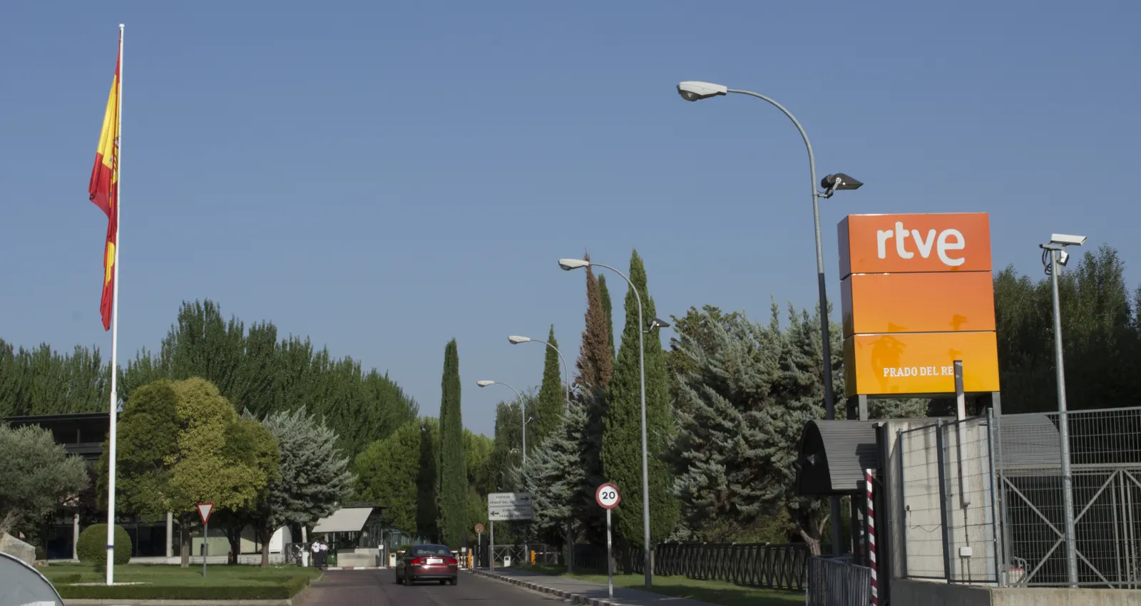 Entrada por carretera a los estudios de RTVE en Prado del Rey, con un cartel naranja con el logotipo «rtve», una bandera de España colgando sin viento de su mástil, arbolado, farolas, cámaras de vigilancia sobre la valla y un coche saliendo del recinto.