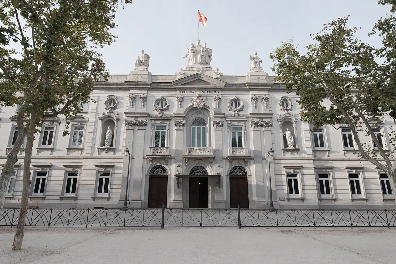 Fachada principal del Tribunal Supremo en Madrid: edificio neoclásico de piedra clara con el rótulo «TRIBUNAL SUPREMO» bajo el frontón, cuatro esculturas de togados sobre la cornisa y la bandera de España ondeando en lo alto. A ambos lados, plátanos de sombra; delante, la explanada vacía tras una verja negra.