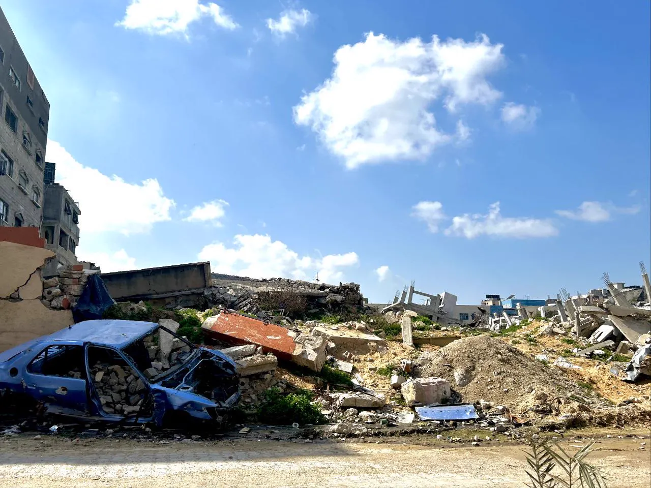 Solar con escombros, un coche calcinado y edificios derruidos en una calle de la Franja de Gaza, bajo un cielo despejado.