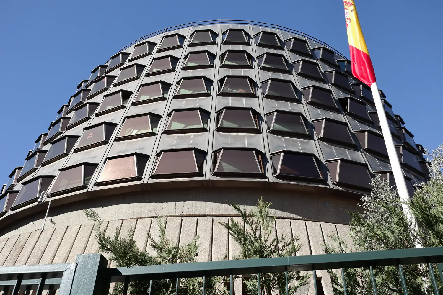 Fachada circular de hormigón del Tribunal Constitucional de España, en Madrid, con sus hileras de ventanas oscuras inclinadas vistas en contrapicado; a la derecha, la bandera española en un mástil, y en primer término una verja verde y varios arbustos.