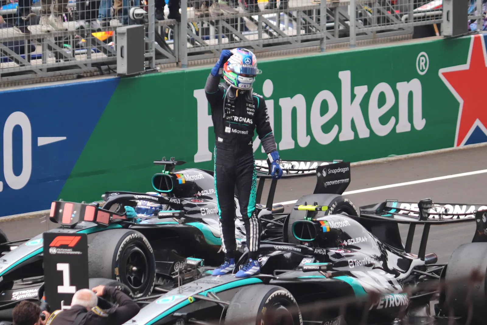Un piloto con mono negro y casco se sube de pie sobre su monoplaza negro de Mercedes, con el puño en alto, junto al cartel con el número 1 del parque cerrado