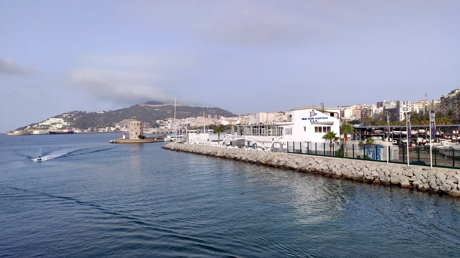 Frente marítimo de Ceuta visto desde el agua: a la derecha, un edificio blanco con el rótulo del Real Club Náutico junto a una escollera de rocas y una hilera de palmeras; detrás, bloques de viviendas de la ciudad; al fondo, a la izquierda, una loma verde con casas dispersas bajo un cielo con nubes bajas.
