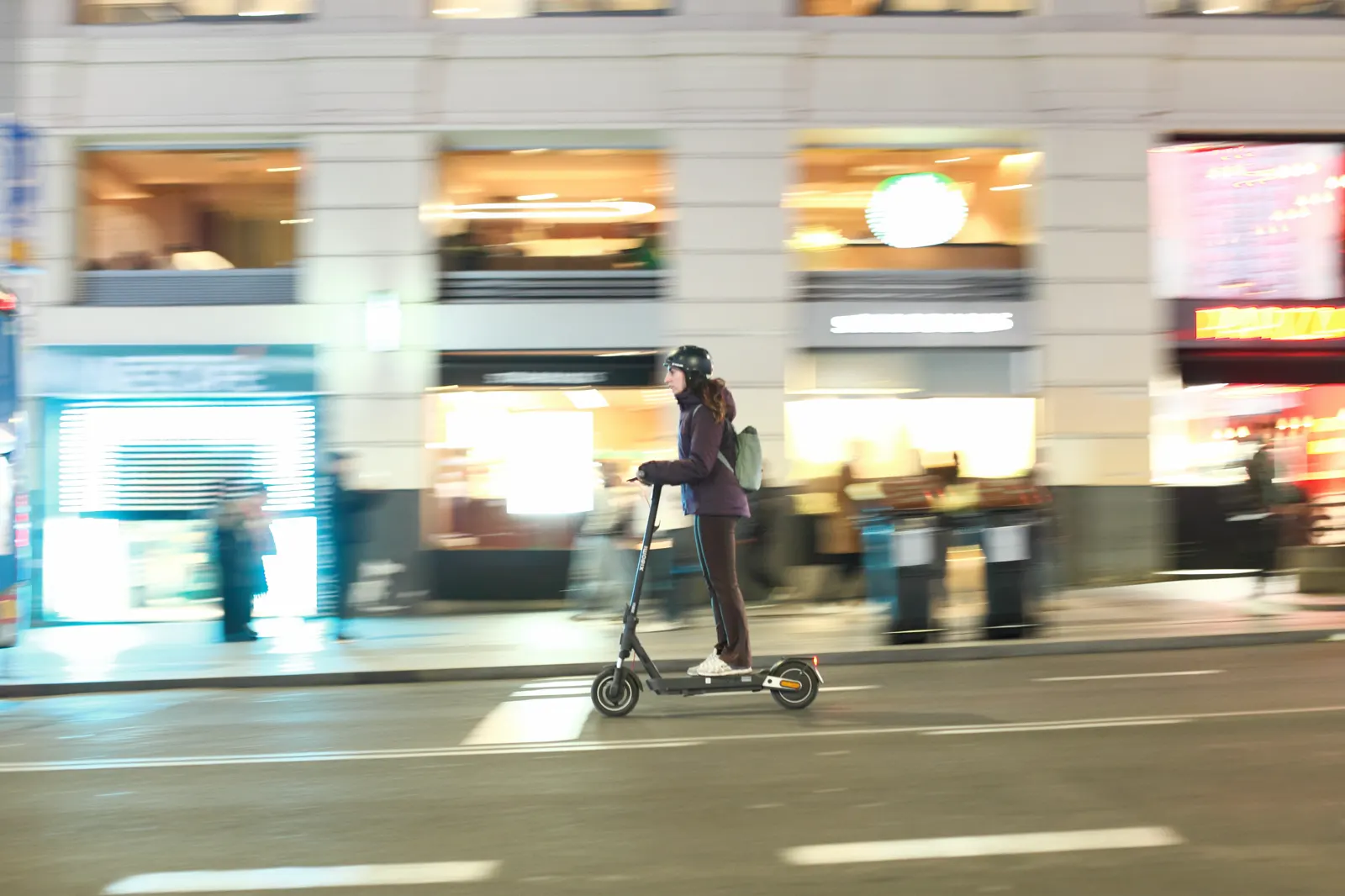 Una persona con casco circula en patinete eléctrico por el carril de la calzada de la Gran Vía de Madrid, de noche. El fondo, con escaparates iluminados y peatones en la acera, aparece movido por el barrido de la cámara.