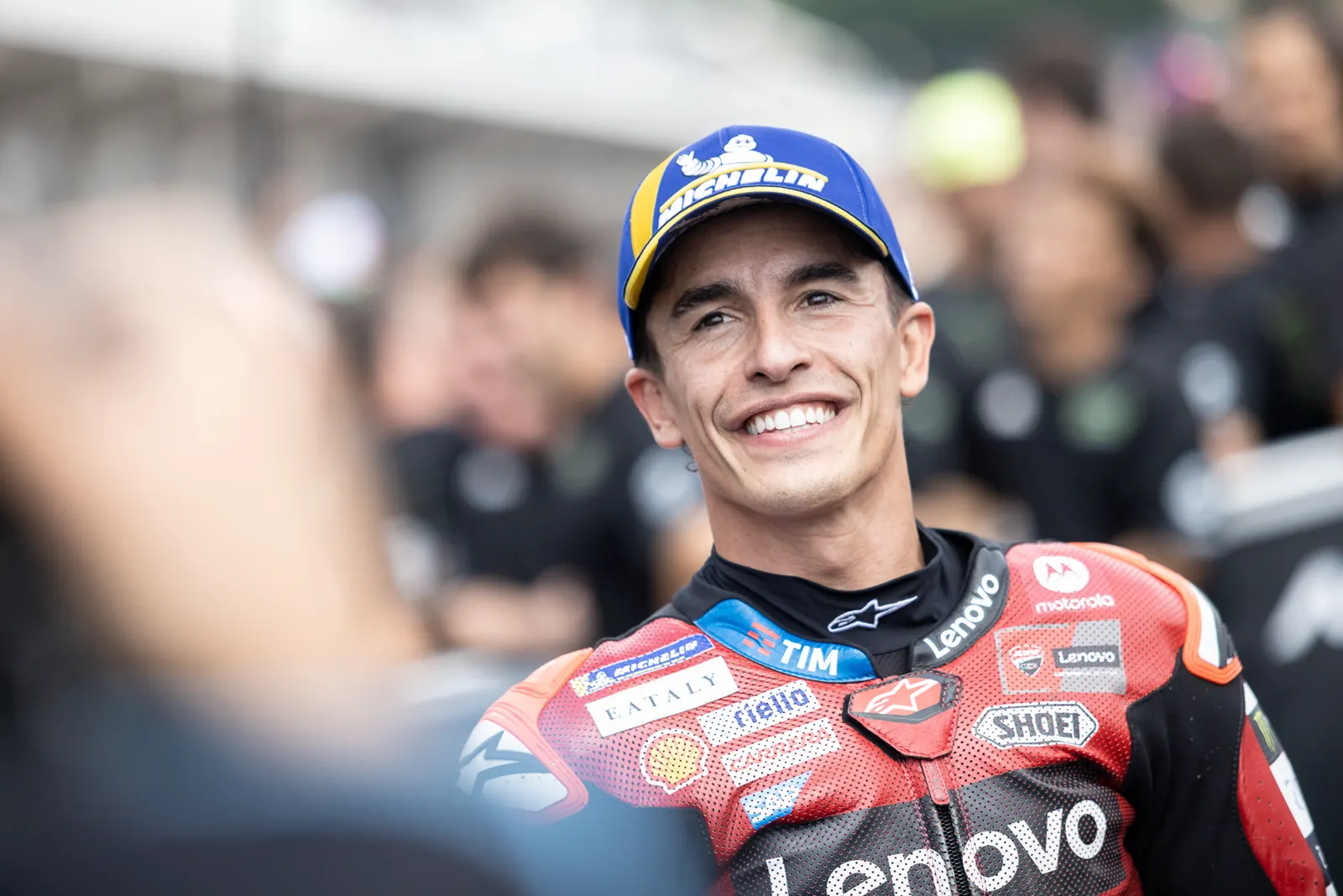 Marc Márquez sonríe en el paddock con el mono de cuero rojo y negro de Ducati Lenovo y una gorra azul de Michelin.