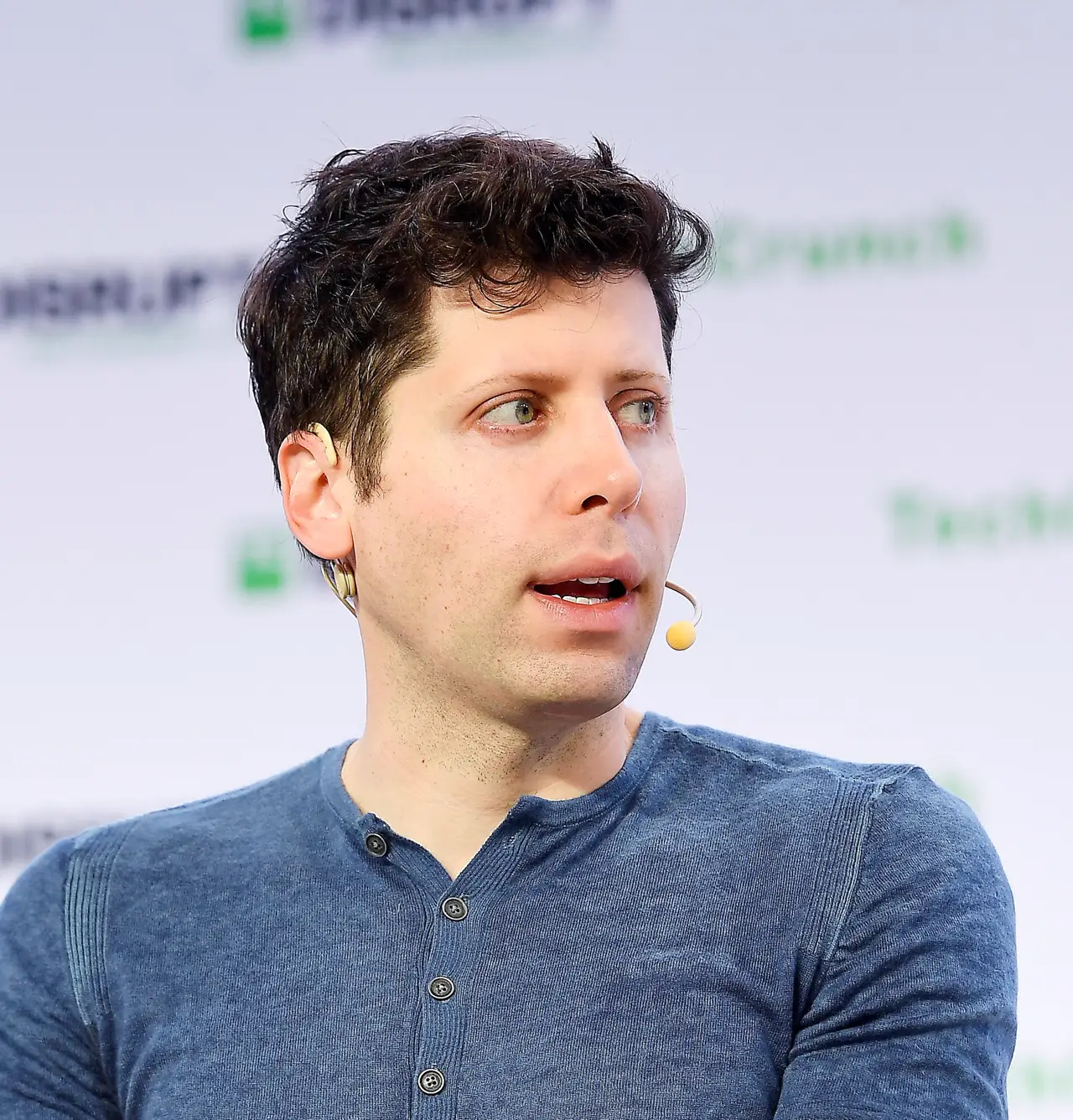 Retrato de Sam Altman, con jersey azul y micrófono de diadema, mirando hacia un lado mientras habla ante un panel con logotipos de TechCrunch Disrupt.