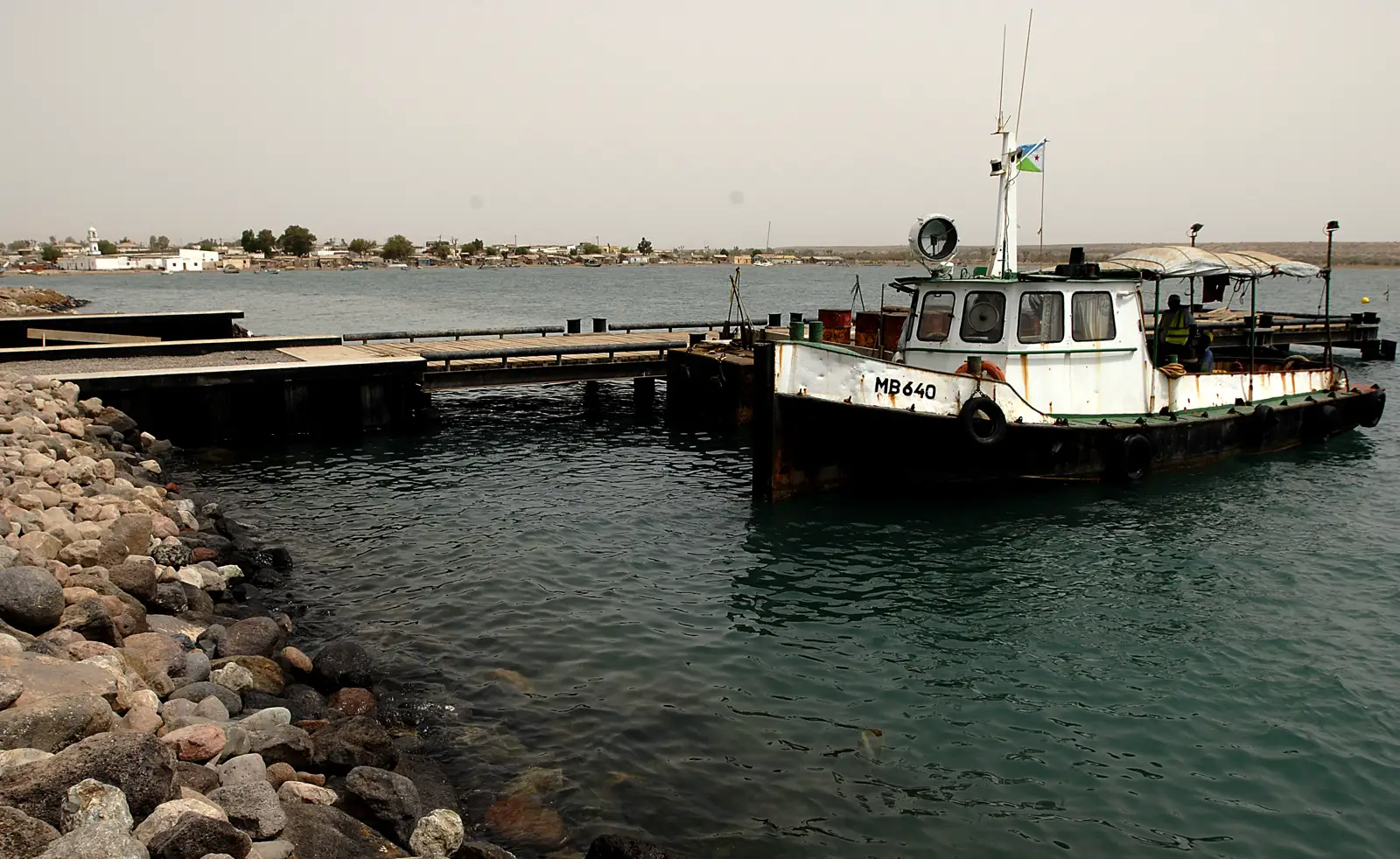 Una pequeña embarcación con bandera de Yibuti amarrada junto al muelle del puerto de Obock, con las casas del pueblo al fondo