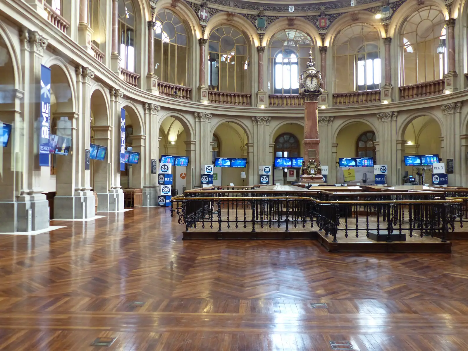 Sala circular del Palacio de la Bolsa de Madrid, con suelo de madera encerada, arcos de piedra y balconadas, una hilera de pantallas encendidas junto a la barandilla del parqué y un reloj sobre una columna al fondo.