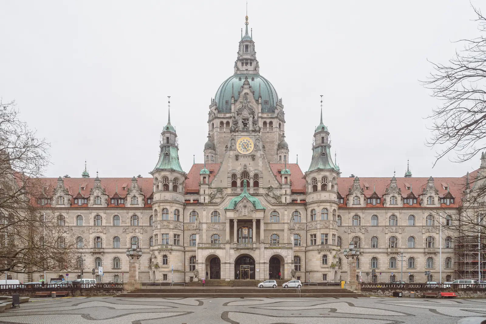 Fachada simétrica de piedra clara del Nuevo Ayuntamiento de Hannover, con cúpula verde de cobre, torres laterales, tejado de teja roja y un gran reloj dorado, vista desde la plaza empedrada que tiene delante en un día nublado de invierno.