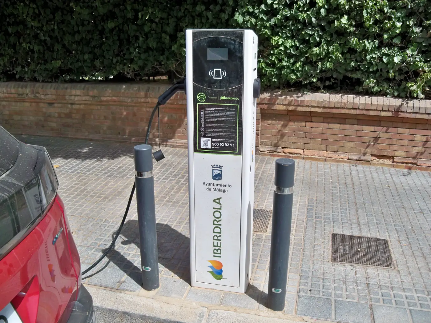 Poste de recarga para coches eléctricos del Ayuntamiento de Málaga e Iberdrola en una acera, con el cable conectado a un vehículo aparcado.