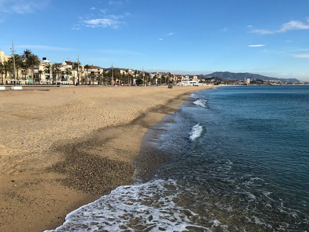 Playa de arena ancha y desierta en primer término, con la orilla batida por espuma baja y el mar azul a la derecha; al fondo, el paseo marítimo de Badalona con palmeras y edificios de pocas plantas y, más allá, la costa y unas montañas bajo un cielo azul casi despejado.