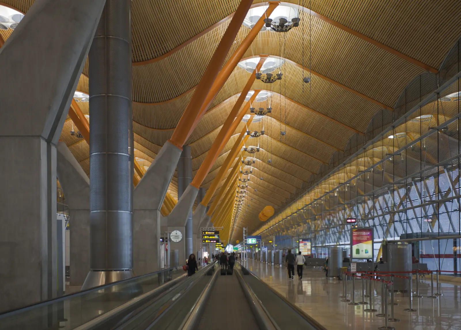 Interior de la terminal T4 del aeropuerto de Madrid-Barajas, con su techo ondulado de bambú sostenido por pilares naranjas y unos pocos pasajeros junto a la cinta transportadora