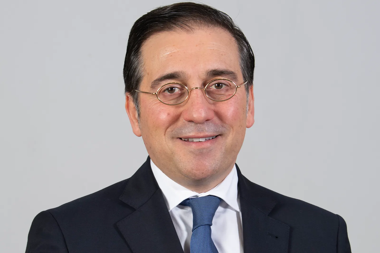 Retrato oficial de José Manuel Albares, ministro español de Asuntos Exteriores: hombre de mediana edad con gafas ovaladas de montura dorada, pelo oscuro peinado hacia atrás, traje azul marino, camisa blanca y corbata azul, sonriente ante un fondo gris liso.